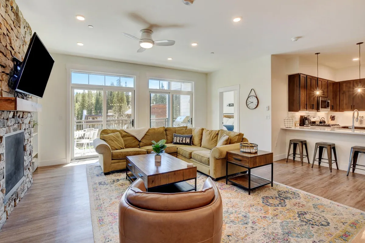A spacious living room anchored by a striking stone fireplace, a cozy sofa, and a kitchen bar area in the background. Large windows let in natural light, creating a warm, inviting atmosphere. The setting evokes family relaxation, game nights, and shared meals.