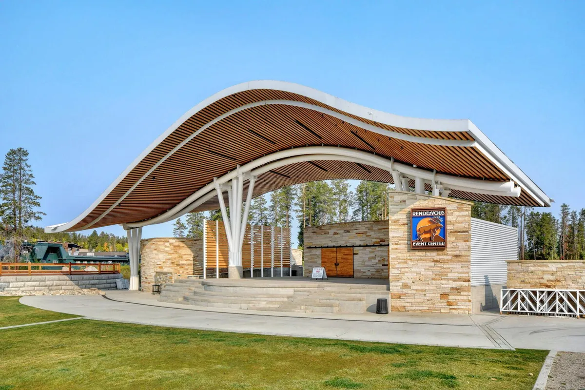 An exterior view of a distinctive curved roof structure with a stone façade, event signage, and open space designed for community gatherings, concerts, and festivals.