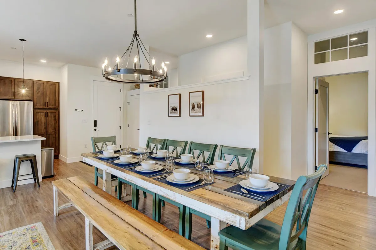 A bright, inviting dining area with a large table set beneath a stylish chandelier, the kitchen visible in the background. Decorative wall art and natural light add warmth and sophistication, making it ideal for festive meals and group activities.