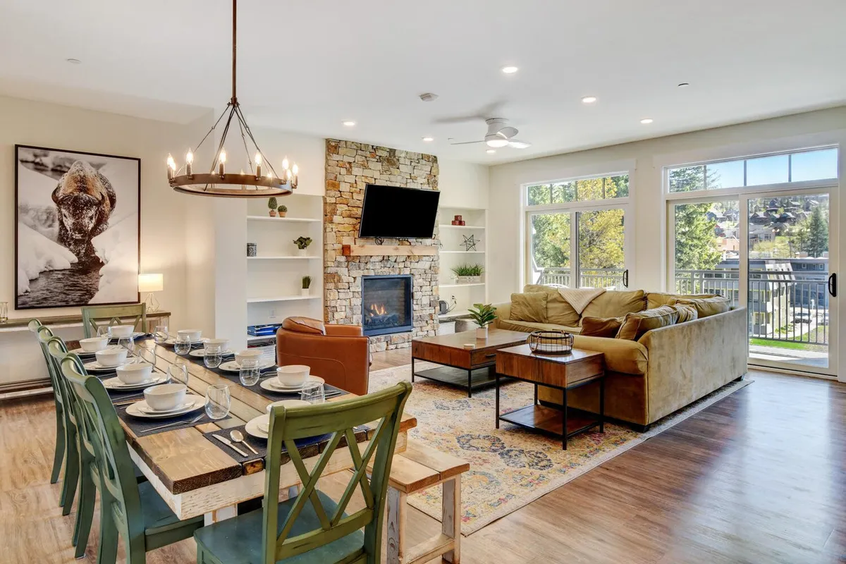 The image showcases a spacious, warmly-lit living and dining area featuring a rustic stone fireplace, a dining table elegantly set for a meal, plush seating perfect for lounging, and expansive windows that frame breathtaking outdoor mountain views. The scene exudes a cozy, inviting atmosphere ideal for family gatherings or unwinding after a day of adventures in Winter Park.