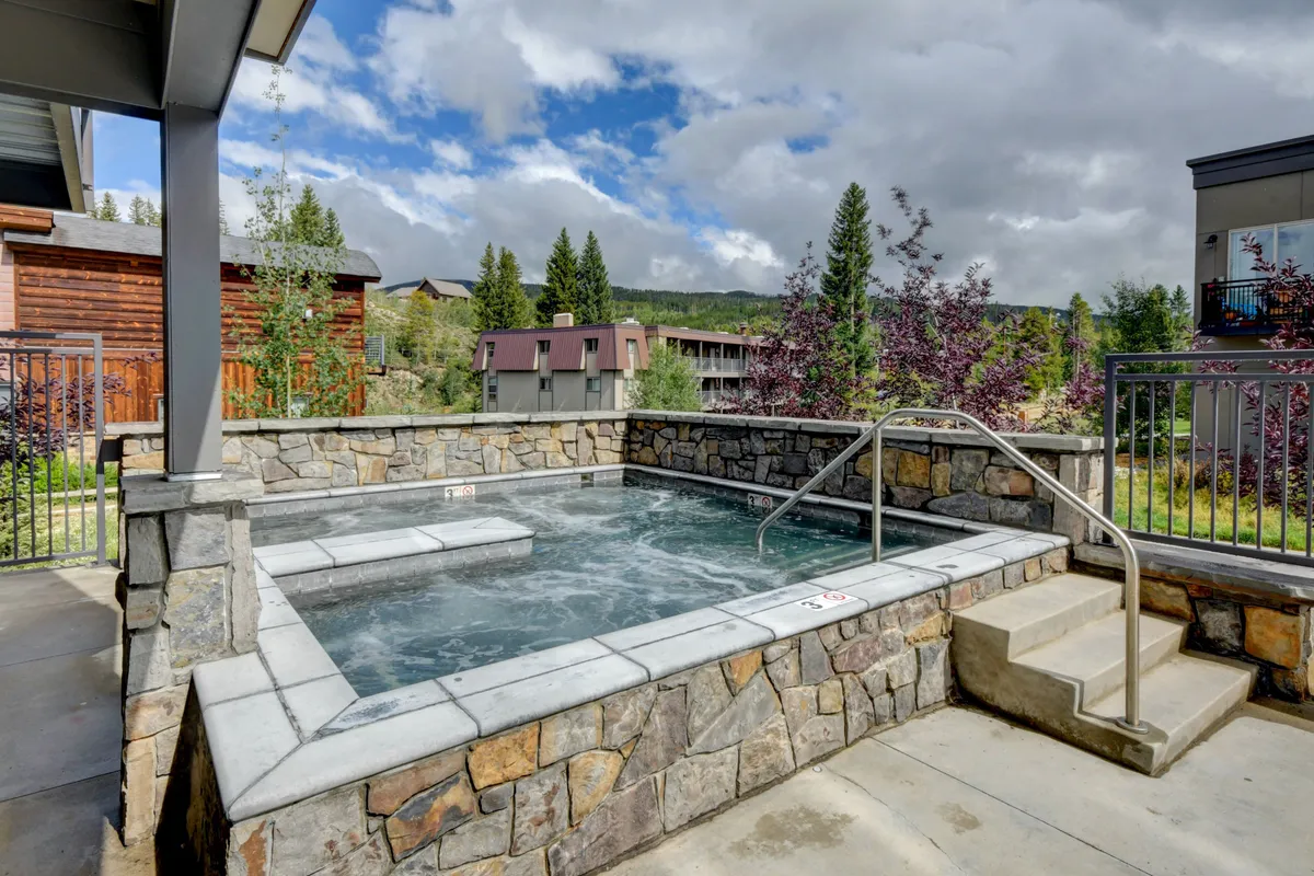 This image depicts a large, inviting hot tub bordered by a rustic stone wall and surrounded by trees, evoking a tranquil and rejuvenating atmosphere. The setting suggests a communal space ideal for relaxing after active days.