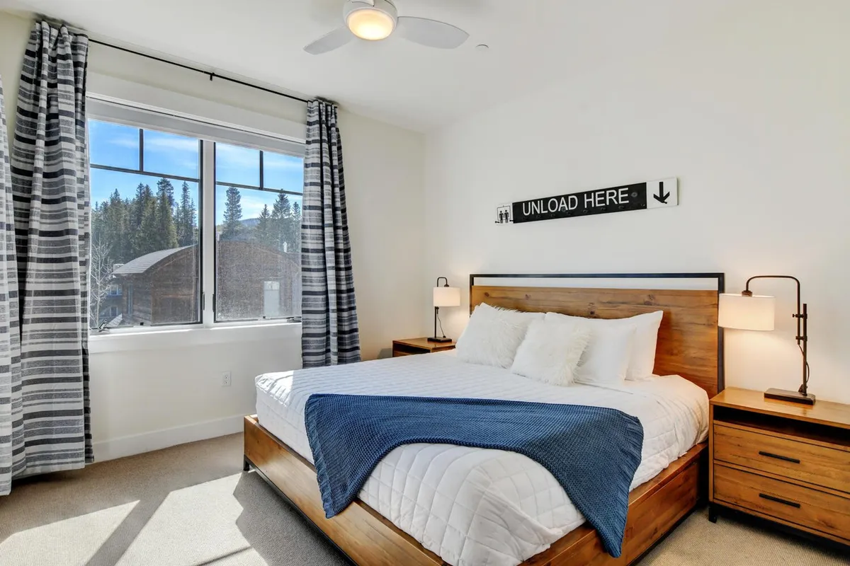 This image shows a bright, inviting bedroom featuring a large bed with crisp white linens, flanked by nightstands and a window with soft curtains. The room is decorated with a calming decorative sign above the bed and enjoys abundant natural light.