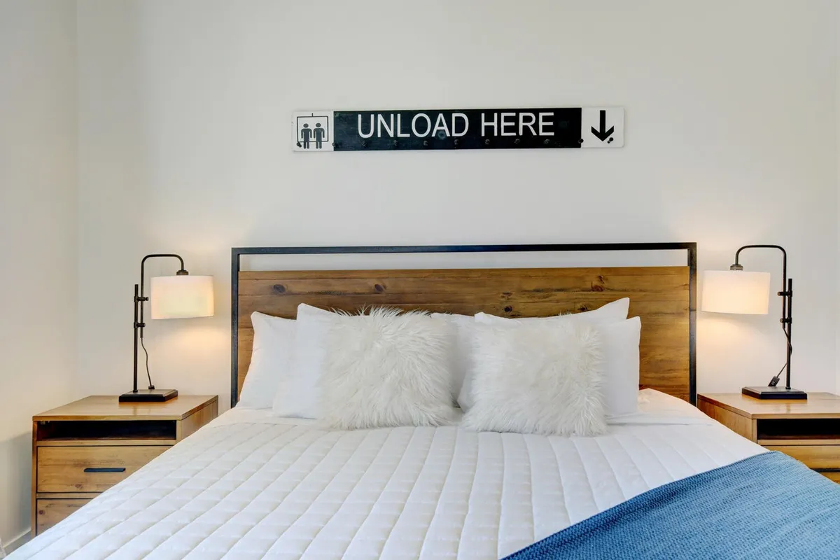 A cozy bedroom with a neatly made bed topped with decorative pillows and soft throws. The room features matching nightstands with lamps and a sign above the bed, radiating warmth and inviting relaxation.