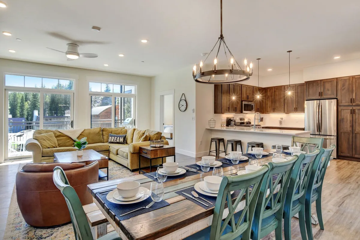 A wide-angle view capturing the integrated living area, dining table, and kitchen island. Plush seating faces large windows that fill the room with light, while the open kitchen with modern appliances and an oversized dining table for 10 invite group gatherings. Hardwood floors and designer lighting set the scene for cozy, communal moments.