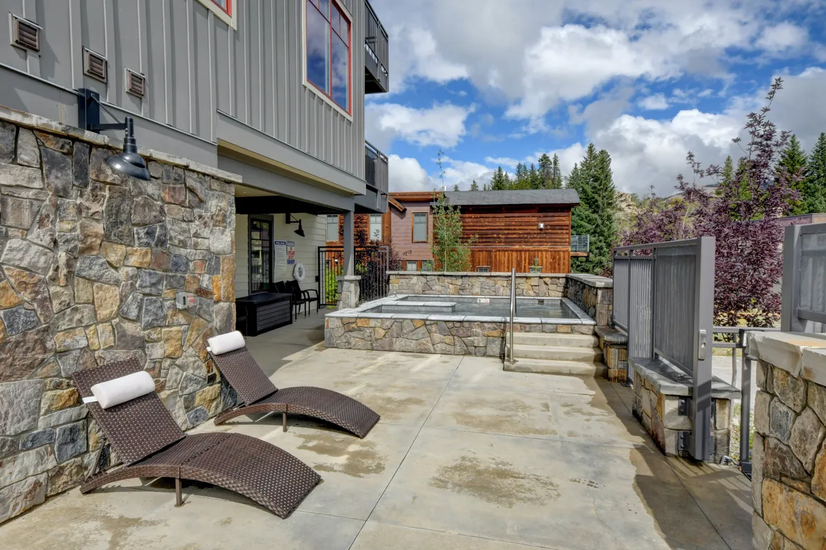A tranquil outdoor patio scene featuring a large stone jacuzzi surrounded by lounge chairs and mature trees. The setting suggests a communal relaxation spot perfect for families and groups to gather after a day of activities.